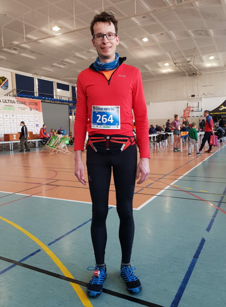 biegacz w czarnych legginsach i czerwonej bluzie stoi sam na sali gimnastycznej, w tle stoliki i ludzie